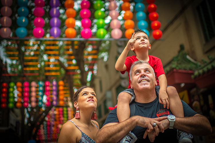 A family enjoying the sights of the Lunar Lanes. Their son is on his fathers shoulders in a Chinatown lane. His mum is looking at something he is pointing at.