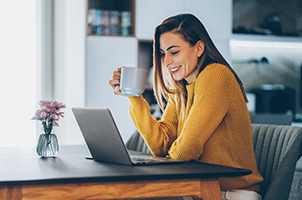A woman looking at her laptop while she is holding a cup of coffee. She is smiling enthusiastically.