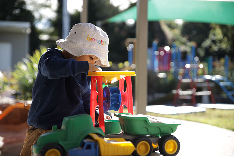 A young child playing with a toy truck. He is wearing a bucket hat with The Benevolent Society logo on it.