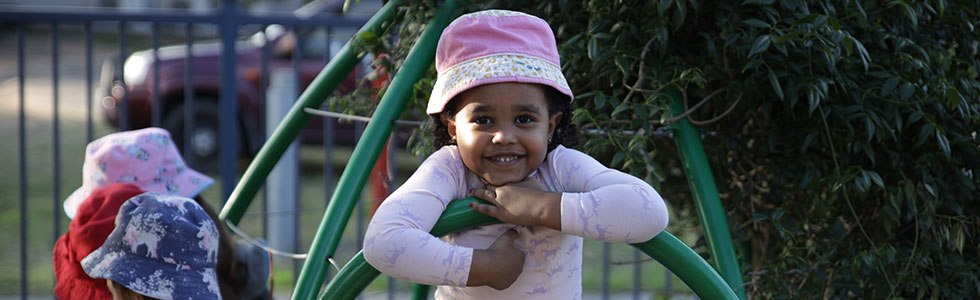 A young girl playing on play equipment outside. She is wearing a hat