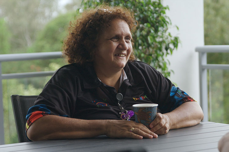 A First Nations employee of The Benevolent Society sitting outside with a cup of coffee. She is wearing a Benevolent Society branded polo shirt with Indigenous patterns on the sleeve