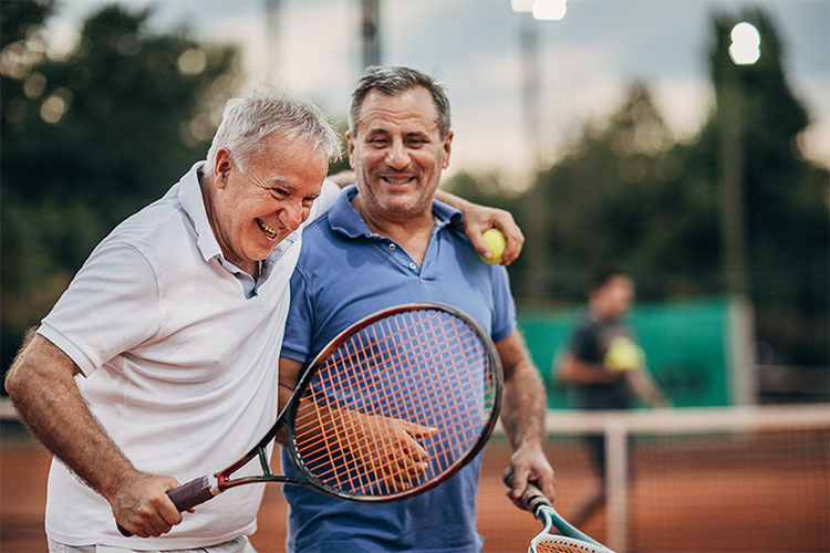 Two older men patting each other on the back after a game of tennis. They are wearing sports attire and holding racquets.