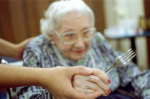 A photo of a senior woman being helped with her fork ready to try some delicious food
