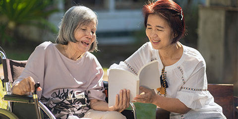 An elderly Asian woman in a wheelchair with a younger Asian woman next to her. They are sitting outdoors.
